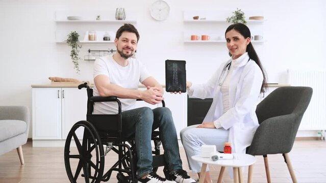 Portrait of young female nurse and mature man wheelchair user, showing x-ray scan of spine on modern tablet. Caucasian woman doctor checking up condition of mature bearded male patient at home.