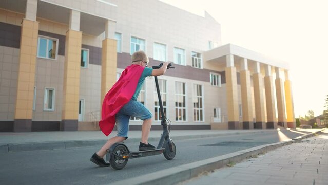 boy superhero on electric scooter. happy family kid lifestyle dream concept. a boy on electric scooter rides on a large square in the city center under a blue sky with clouds sun. superhero boy