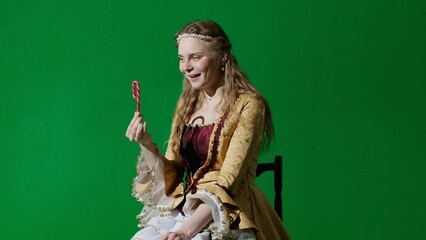 Woman in ancient outfit on the chroma key green screen background. Female in renaissance style dress sitting holding candy cane eating sweets enjoying.