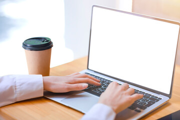 Female hands in white shirt typing on laptop, white screen, empty space. Close-up photo of hands, laptop on wooden table, glass with hot drink, business, freelancing, study