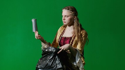 Woman in ancient outfit on the chroma key green screen background. Female in renaissance dress holding garbage bag looks inside takes out trash cans.