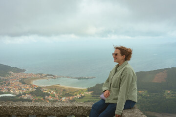 Young woman sitting on a viewpoint overlooking the village of Cariño, Ortigueira.