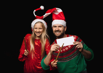 A young male music teacher teaches a girl in a red dress and Christmas hats to play the drums, the girl holds drumsticks in her hands, the concept of a music lesson in the studio on a black background