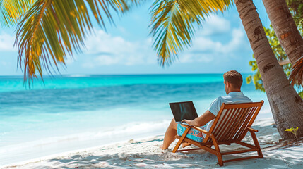 A man sitting on a deck chair with a laptop on a sandy beach, palm trees around, facing the ocean. ...