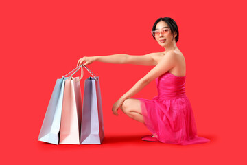 Happy beautiful young Asian woman with shopping bags sitting on red background