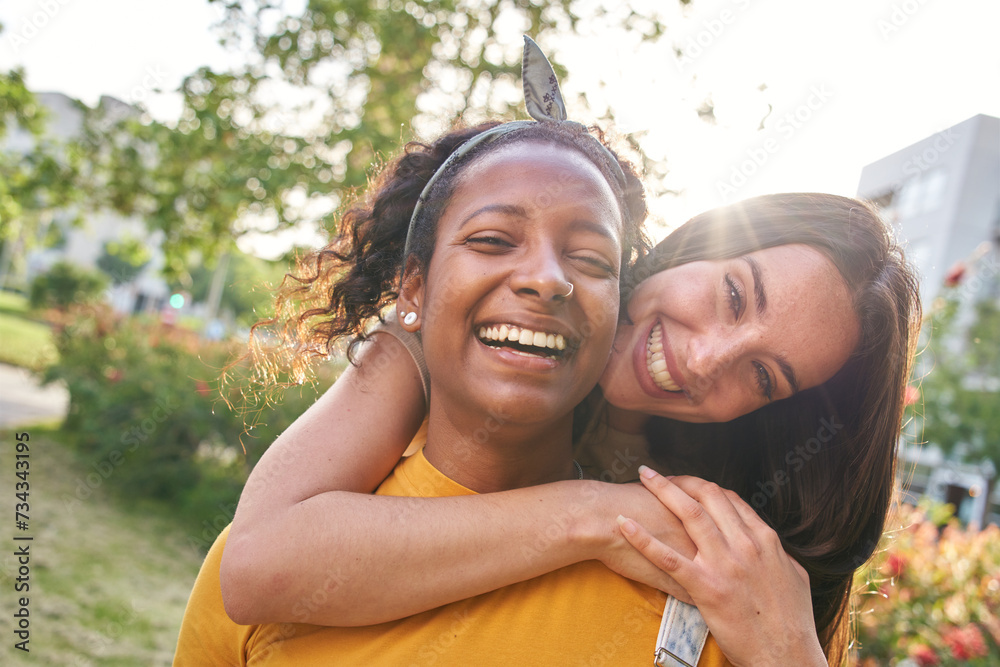 Wall mural Portrait young friends happy in park flare lens in background. Attractive Caucasian woman hugs smiling latina girl in affectionate attitude. Multiracial females looking at cheerful camera outdoor.