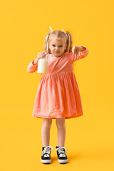 Displeased little girl with bottle of milk showing thumb-down on yellow background