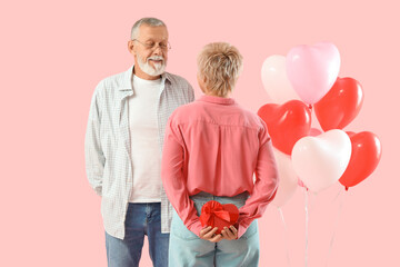 Mature woman hiding gift from her husband behind back on pink background. Valentine's Day celebration