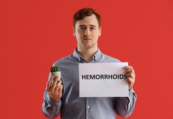 Young man holding paper with word HEMORRHOIDS and cactus on red background