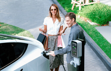 Young couple travel with EV electric car charging in green sustainable city outdoor garden in...