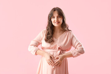 Young pregnant woman showing heart gesture on pink background. Valentine's Day celebration