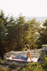 Fit young woman practices yoga on the rocks against the background of the pine forest. A beautiful girl meditates and does breathing exercises in the mountains.