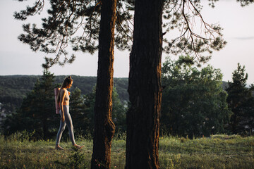 Fototapeta premium Happy young woman in fitness clothes with yoga mat going to workout in the forest at sunset.