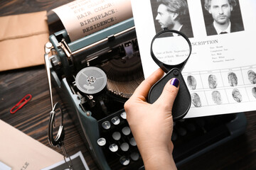 Female hand with magnifier, retro typewriter and wanted poster on wooden background, closeup