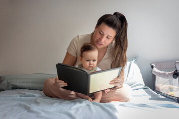 Madre sentada en su cama con su bebé varón leyéndole un cuento en un día soleado