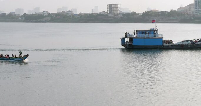 A ferry full of passengers departs,  a small motorized pirogue brimming with people sails along the riverbank.