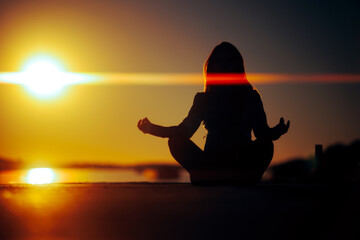 Silhouette of a Woman Sitting in a Lotus Pose Looking at the Sunset. Mindful carefree person relaxing outdoors enjoying he morning 
