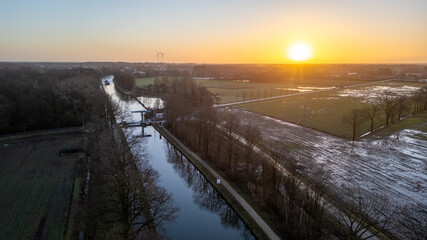 This aerial photograph captures a crisp winter sunrise, with the sun hovering just above the horizon, casting a soft golden light across the landscape. The canal running through the scene reflects the