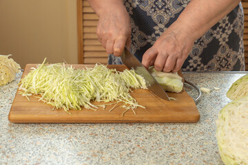Woman thinly slices white cabbage with a knife on a wooden cutting board to prepare a vegetable...