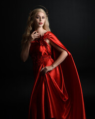 Fototapeta premium Close up portrait of beautiful blonde model wearing flowing red silk toga gown and crown, dressed as ancient mythological fantasy goddess. Graceful elegant pose isolated on dark studio background.