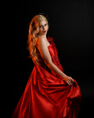Fototapeta premium Close up portrait of beautiful blonde model wearing flowing red silk toga gown and crown, dressed as ancient mythological fantasy goddess. Graceful elegant pose isolated on dark studio background.