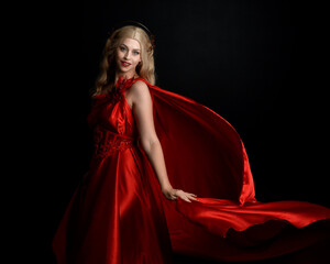 Fototapeta premium Close up portrait of beautiful blonde model wearing flowing red silk toga gown and crown, dressed as ancient mythological fantasy goddess. Graceful elegant pose isolated on dark studio background.