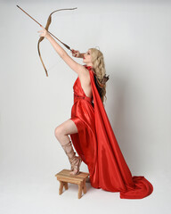 Full length portrait of  blonde model dressed as mythological fantasy goddess in flowing red silk toga gown, crown. Graceful elegant pose  holding  archery weapons, isolated  on studio background
