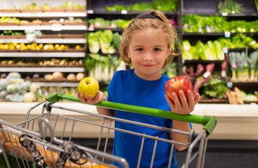 Kid with apple and shopping cart at grocery store. Funny cute child on shopping in supermarket. Grocery store. Grocery shopping, healthy lifestyle concept.