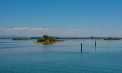 An island in the Grado section of the Marano and Grado Lagoon in Friuli-Venezia Giulia, N.E. Italy. Channel markers shows the edge of a navigable channel. Grado town is in the background - obrazy, fototapety, plakaty