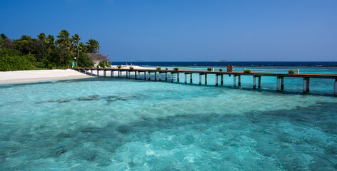 Wooden bridge over the sea leading to an island with sandy beaches and a green forest. Palm trees.