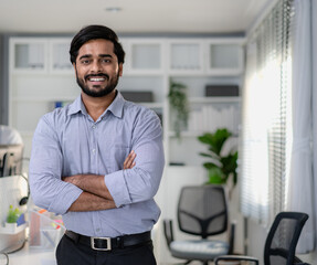 Friendly Indian businessman standing arms crossed in modern office smiling. Confident young smart bearded man entrepreneur looking at camera smiling. Successful India ethnic manager work in workplace.