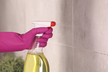 Woman spraying detergent on gray tiled wall indoors, closeup. Space for text