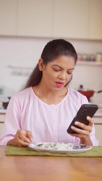 vertical shot of Indian young woman using mobile phone while eating food at home - concept of social media addiction, cyberspace and surfing internet