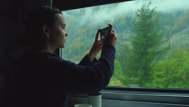 Happy woman taking pictures of Carpathian mountains from train. Girl takes photo of mountain landscape outside the window while sitting inside transport. Passenger or traveler with mobile phone