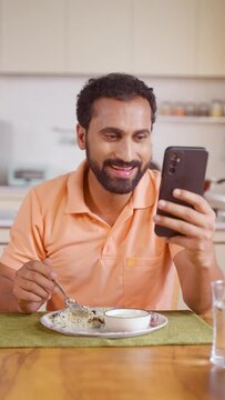 Vertical shot of Indian middle aged man busy using mobile phone while eating lunch on dining table at home - concept of modern lifestyles, internet distraction and social media sharing