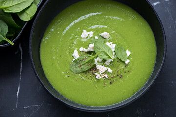 Bowl of spinach cream-soup topped with feta cheese, horizontal shot on a black marble surface, middle close-up