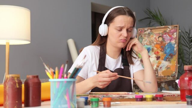 Sad Caucasian creative woman painter in apron painting in her studio abstract picture and listening to music in headphones having not inspire to drawing