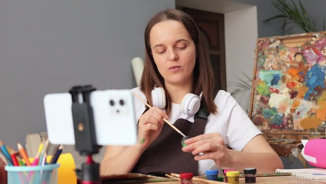 Adorable Caucasian brown haired woman wearing apron sitting at workplace using cell phone on tripod for recording online lessons of creating works of art in her studio
