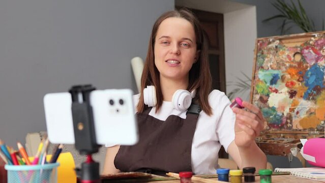Professional female painter shooting video tutoring of painting creating online course of watercolor paint Caucasian brown haired woman wearing apron painting at her workplace in art studio