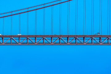 Part of the 25 de Abril suspension bridge with red steel cables that support the structure where cars, vans and trucks circulate to access Lisbon, under a clear blue sky.