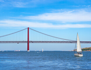 Obraz premium Motorboats, container ship, and sailboats sailing on the Tagus River with the red steel 25 de Abril suspension bridge in the background with road traffic accessing Lisbon, under a clear blue sky.