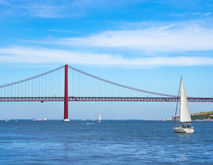 Motorboats, container ship, and sailboats sailing on the Tagus River with the red steel 25 de Abril suspension bridge in the background with road traffic accessing Lisbon, under a clear blue sky.
