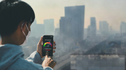 Asian man wearing the N95 using a smartphone app showing PM 2.5 dust in the capital that exceeds the standard that is harmful to health. air pollution concept, pm 2.5 warning.health care, environment.