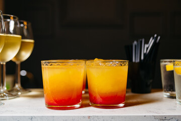 Tequila Sunrise Cocktails and White Wine Glasses. Two vibrant Tequila Sunrise cocktails with a gradient of orange and red, beside glasses of white wine on a bar counter.