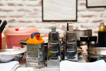 Professional Kitchen Graters and Cooking Equipment. A busy professional kitchen workspace featuring stainless steel graters and various cooking utensils, with a brick wall in the background.
