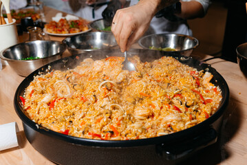 Chef Preparing Seafood Paella in Large Pan. A chef's hand stirs a large pan of seafood paella, rich with rice, shrimp, and mussels, a traditional Spanish dish.