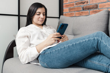 Beautiful young woman lying on a couch and using mobile phone, smartphone for social media, internet browsing or messaging