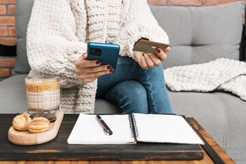 Unrecognizable woman sitting on a couch, holding credit card and using mobile phone or smartphone. glass of coffee drink with dessert and notepad with a pen on the table - obrazy, fototapety, plakaty