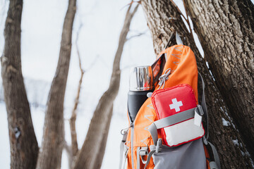 Orange backpack with a first aid kit and a thermos bottle hanging on a tree in the winter forest.