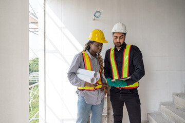 Two construction workers checking work schedule on tablet computer. Wear hardhat and safety vest, stand between stairway in construction site.